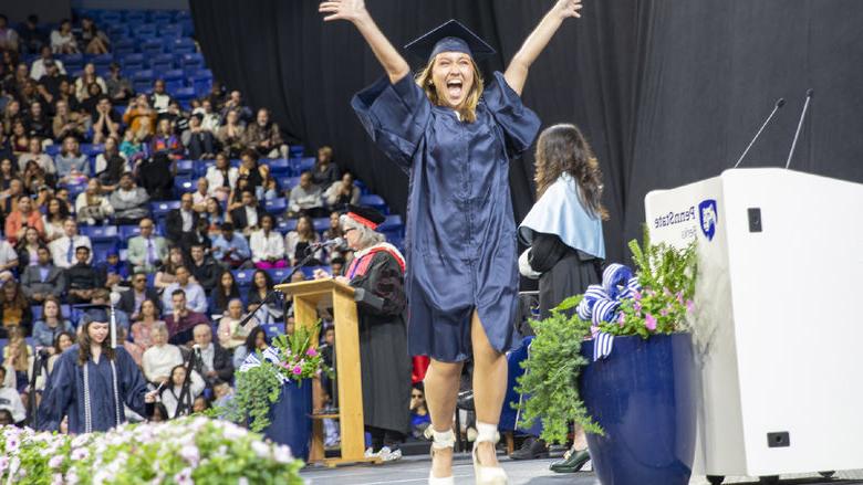 Student wearing cap and gown jumps for joy crossing the stage at commencement