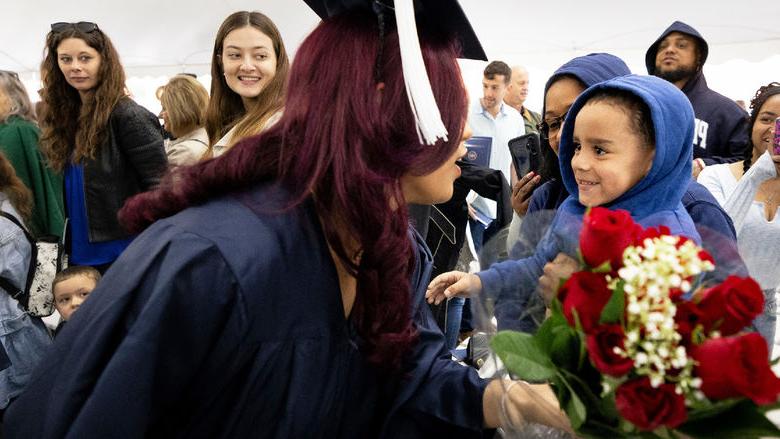 Woman in commencement gown bends down to accept a bouquet of flowers from a young boy.