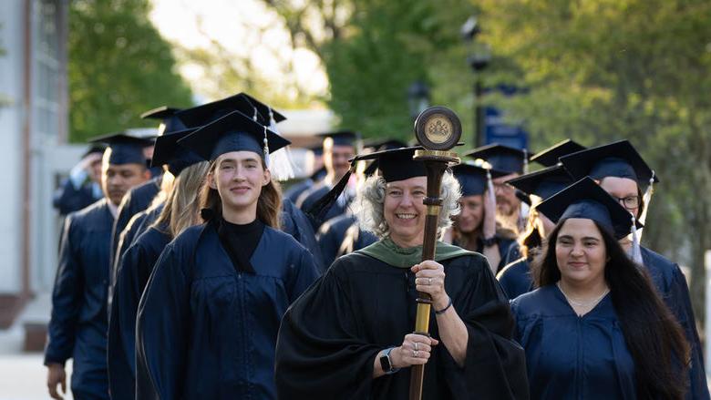 Graduates in caps and gowns in two rows walking down sidewalk led by faculty member in black robe carrying a staff and pendant.